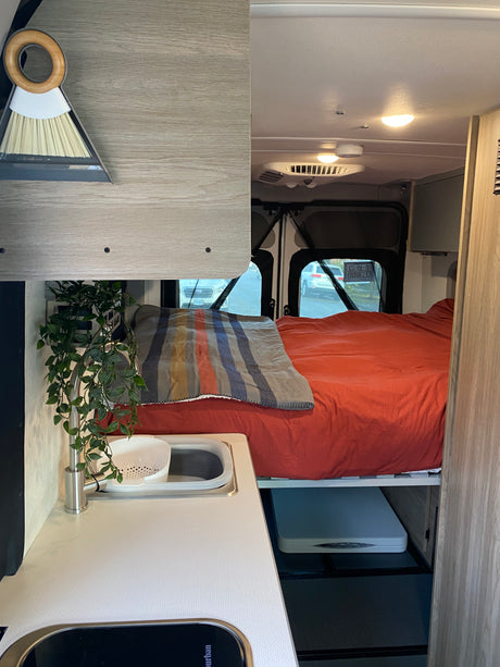 Inside an rv rental: a light wood-themed camper van interior with a bed, small sink, and overhead storage.