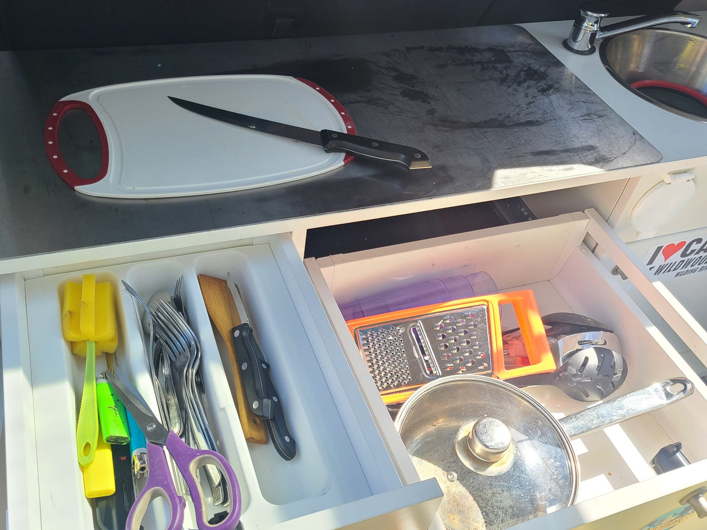 Compact RV rental kitchen setup: organized drawers with utensils, knives, grater, pot, plus sink & cutting board. — photo 3