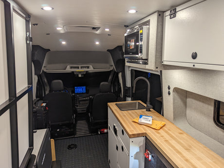 Inside an RV rental: a compact kitchen area with wood countertop, sink, microwave, and driver's cabin visible forward.