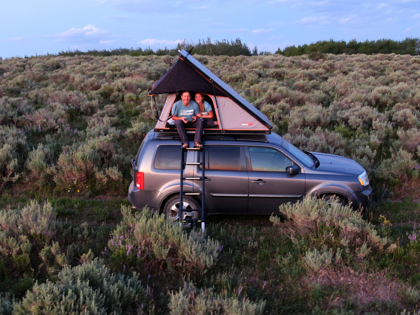 Happy couple enjoys rooftop tent camping on their SUV, a compact alternative to an RV rental for a nature escape.