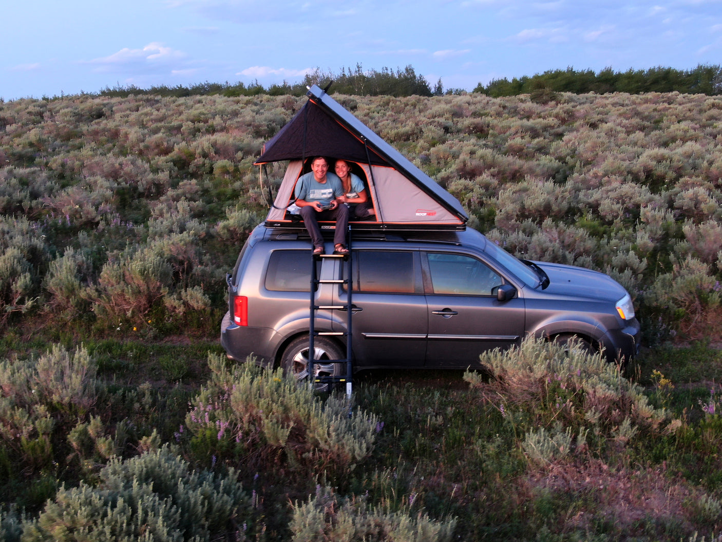 Happy couple enjoys rooftop tent camping on their SUV, a compact alternative to an RV rental for a nature escape.