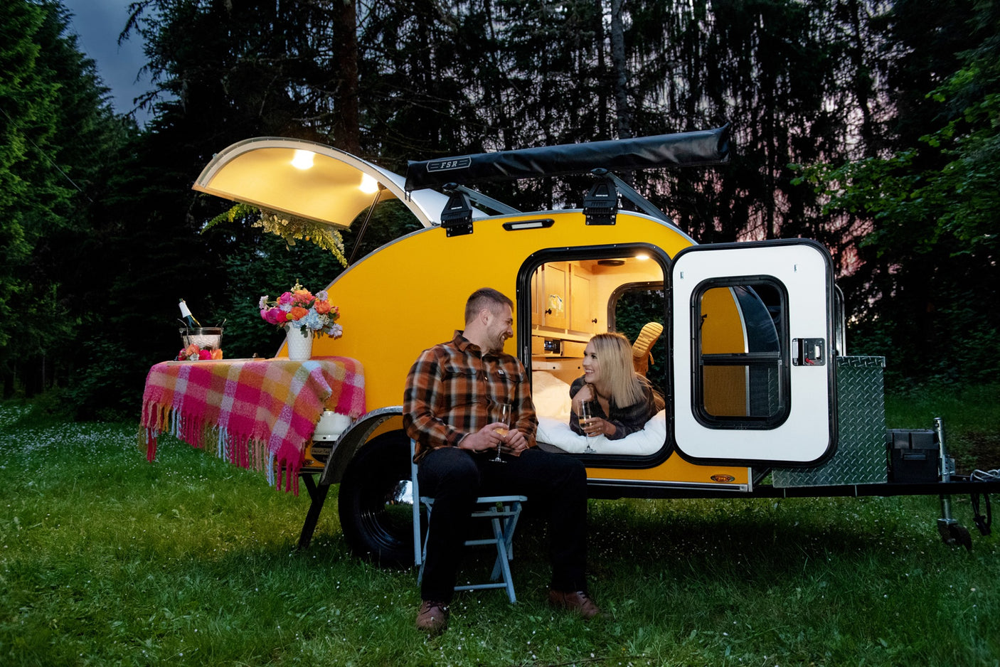 Cozy RV rental adventure: A couple enjoys champagne by their yellow teardrop camper at dusk, ideal for glamping.