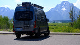An RV rental: a gray Winnebago Revel camper van with roof ladder, backed onto asphalt, offering views of majestic mountains.