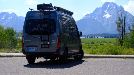 An RV rental: a gray Winnebago Revel camper van with roof ladder, backed onto asphalt, offering views of majestic mountains.