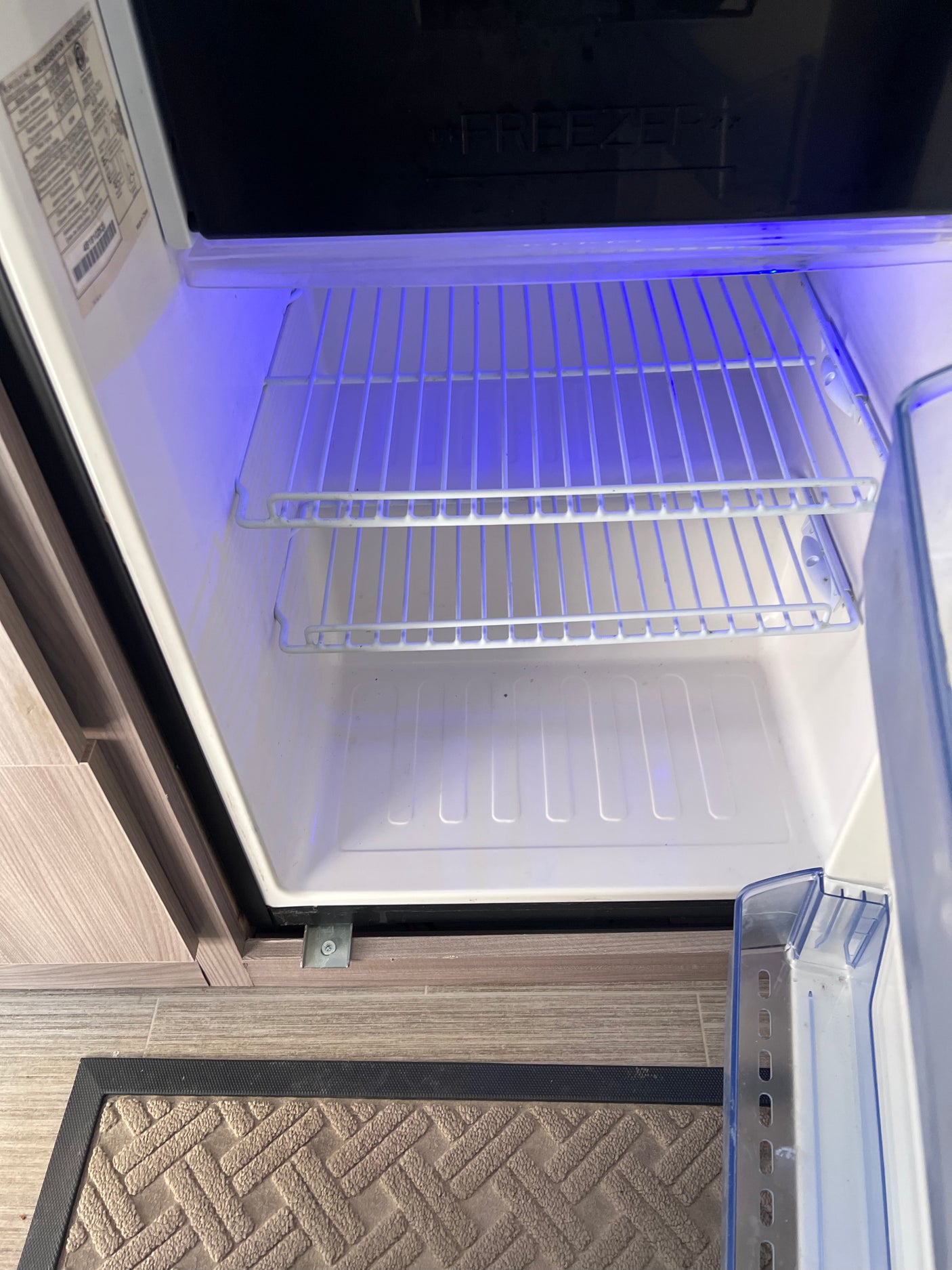 RV rental kitchen: empty mini-fridge interior with blue light, visible freezer, and wire shelves, ready for use.