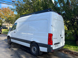 White Mercedes Sprinter van with a roof rack, commonly used for RV rental, parked on a suburban street.