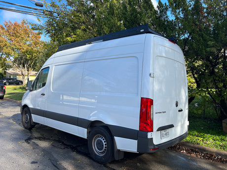 White Mercedes Sprinter van with a roof rack, commonly used for RV rental, parked on a suburban street.