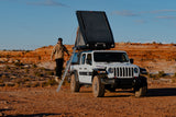 This 4x4 Jeep with a rooftop tent is an ideal RV rental for desert overlanding. Man on ladder, red rock backdrop.