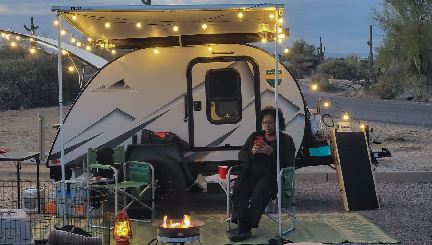 Enjoying an RV rental adventure! A woman relaxes by a fire pit under string lights next to her travel trailer in the desert.