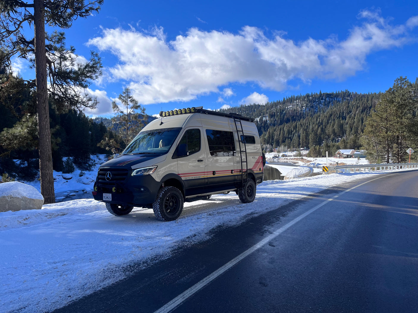 A beige Storyteller Overland rv rental van parked on a snowy mountain road, next to a partially frozen river.