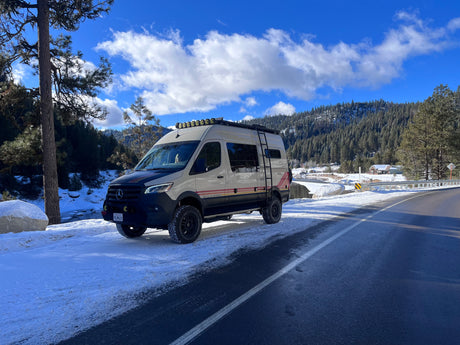 A beige Storyteller Overland rv rental van parked on a snowy mountain road, next to a partially frozen river.