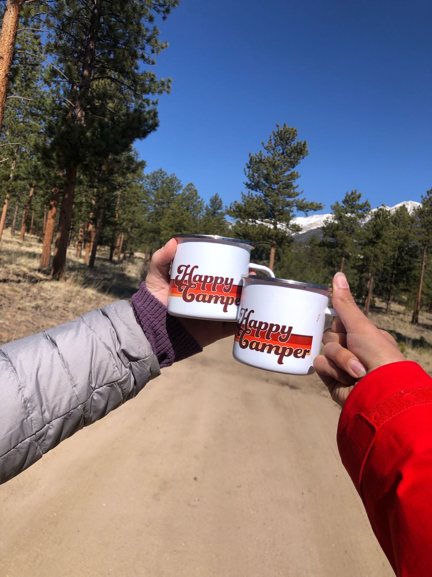 Hands holding 'Happy Camper' mugs on an RV rental adventure. Forest path, pine trees, and snowy peaks under blue sky.