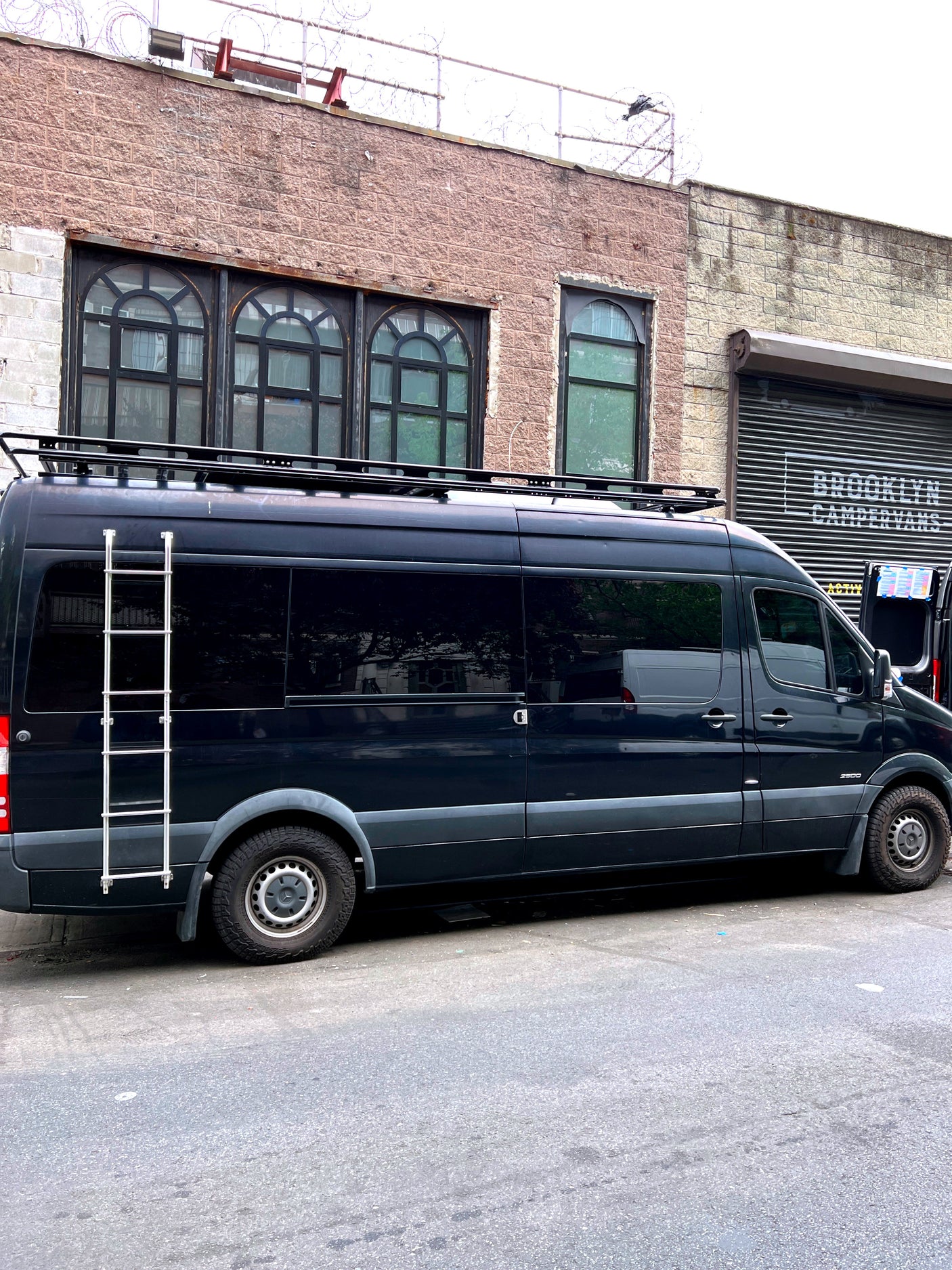 Black Mercedes Sprinter RV rental with roof rack and ladder, parked at Brooklyn Campervans, ready for adventure.