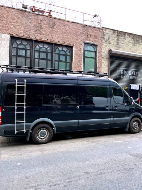 Black Mercedes Sprinter RV rental with roof rack and ladder, parked at Brooklyn Campervans, ready for adventure.