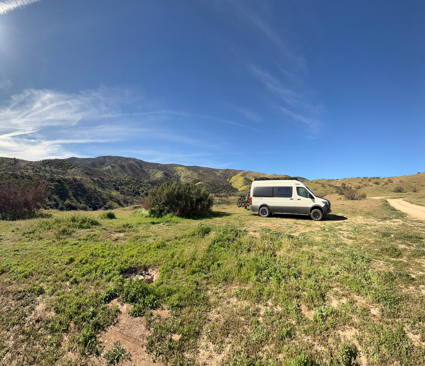 White RV rental camper van with bikes on rear, parked in a sunny, green hilly landscape under a clear blue sky.