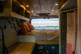 Cozy rv rental interior: a bed with rust-orange pillows faces a large window with a serene lake view and boat.