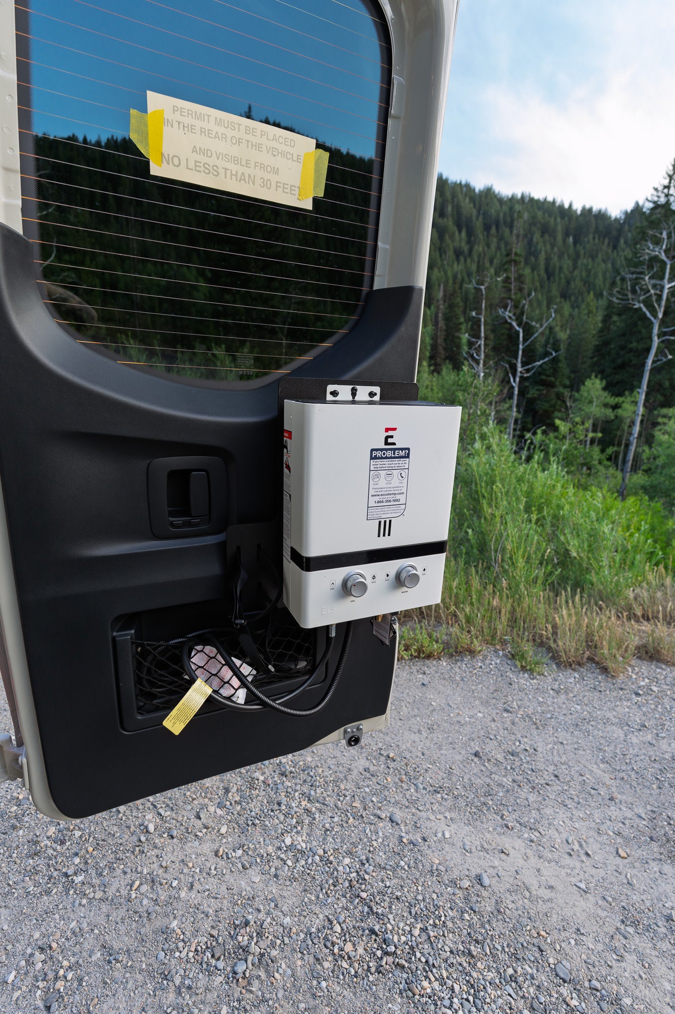 An RV rental's open rear door reveals a mounted portable water heater, ideal for outdoor showers, plus a rear visibility permit.