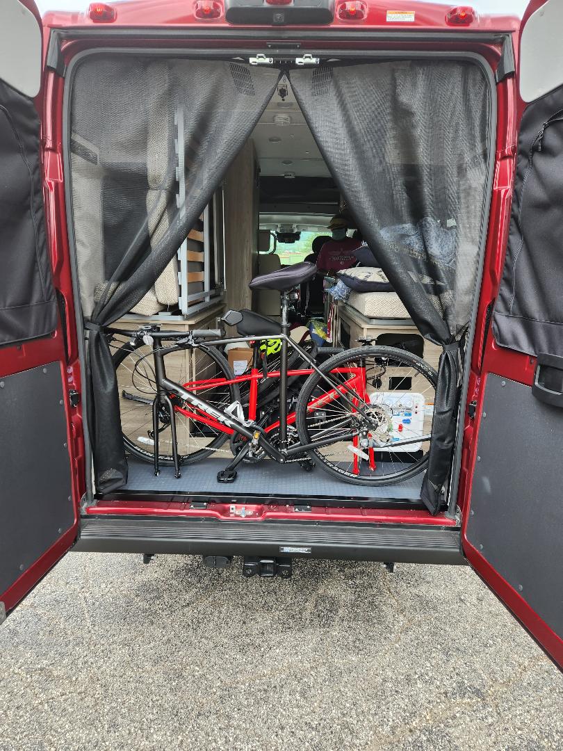 Red RV rental with open rear doors, showcasing interior bike storage and living area. Mesh screens visible for a camper van.