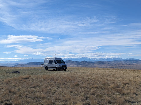 RV rental for remote adventures: A grey camper van on a vast, dry field with distant snowy mountains & blue sky.