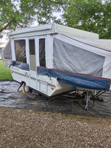 White Rockwood pop-up camper set up on gravel under trees. Ideal for an RV rental adventure.