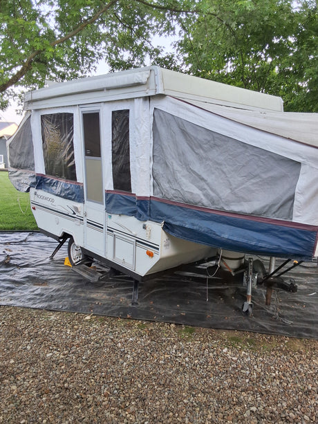 White Rockwood pop-up camper set up on gravel under trees. Ideal for an RV rental adventure.