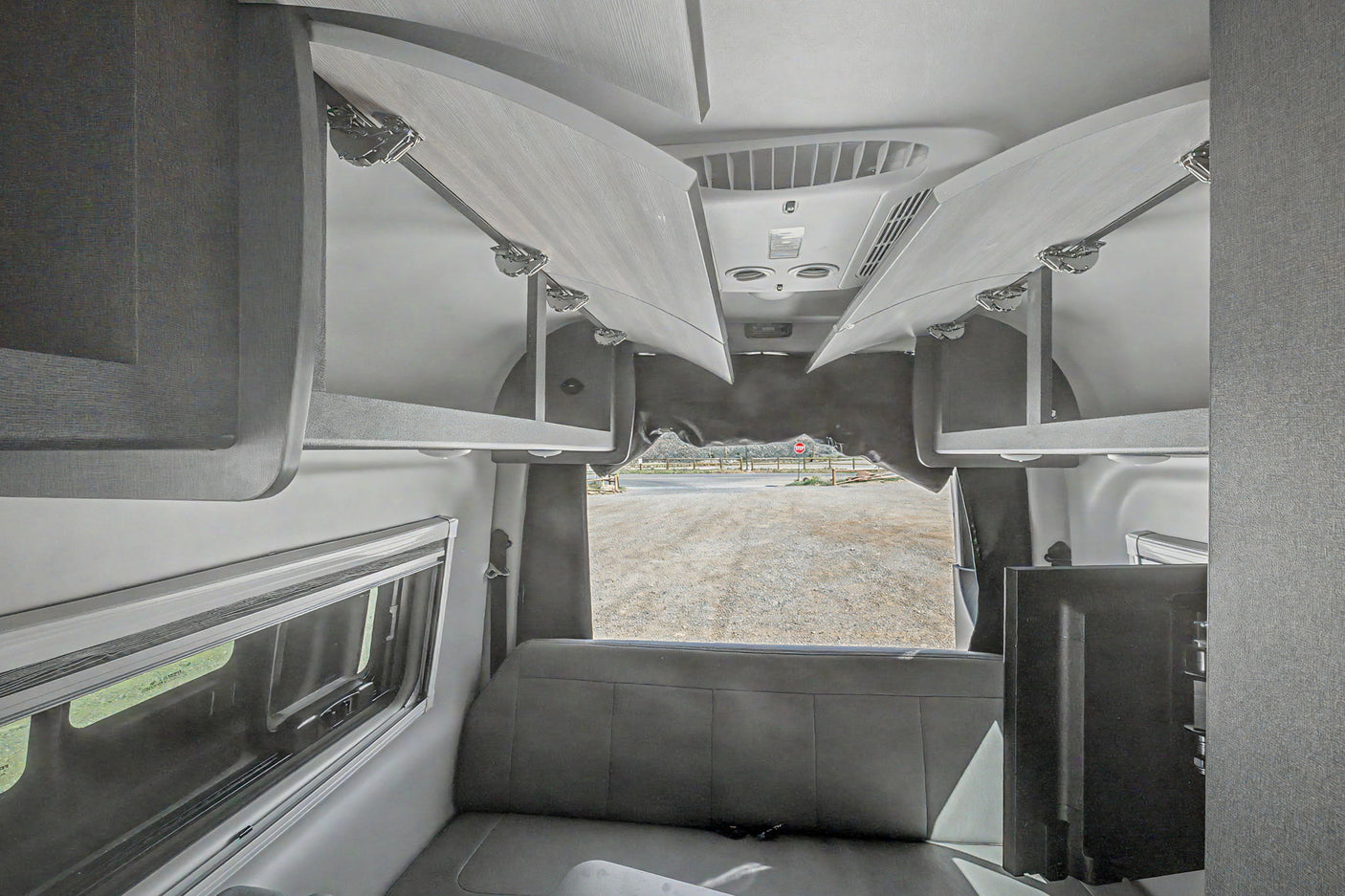 Spacious RV rental interior featuring open overhead storage, bench seating, a window, and a TV, with a view of a gravel lot.