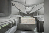 Spacious RV rental interior featuring open overhead storage, bench seating, a window, and a TV, with a view of a gravel lot.