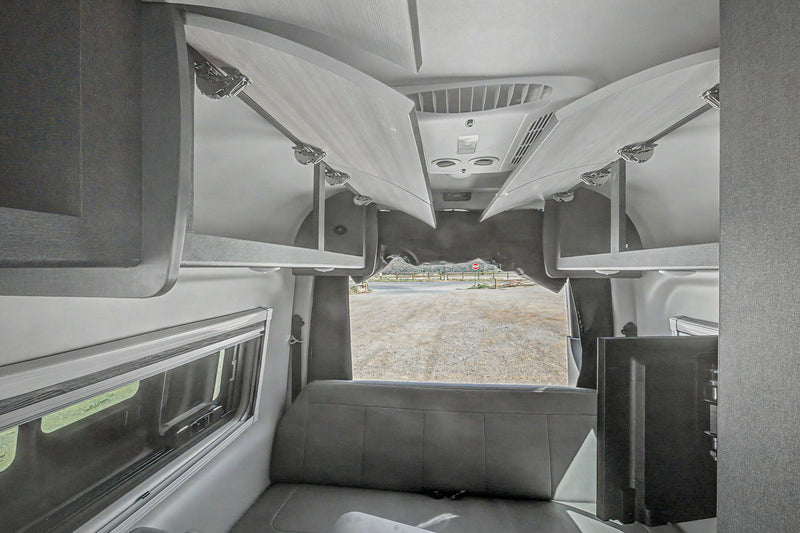Spacious RV rental interior featuring open overhead storage, bench seating, a window, and a TV, with a view of a gravel lot. — photo 3