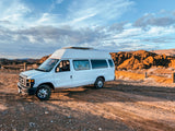 White van with high top, like an RV rental, parked on a dirt road in arid mountains under a dramatic sky. Person inside.
