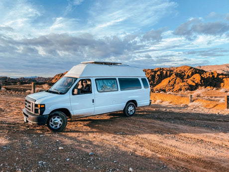 White van with high top, like an RV rental, parked on a dirt road in arid mountains under a dramatic sky. Person inside.