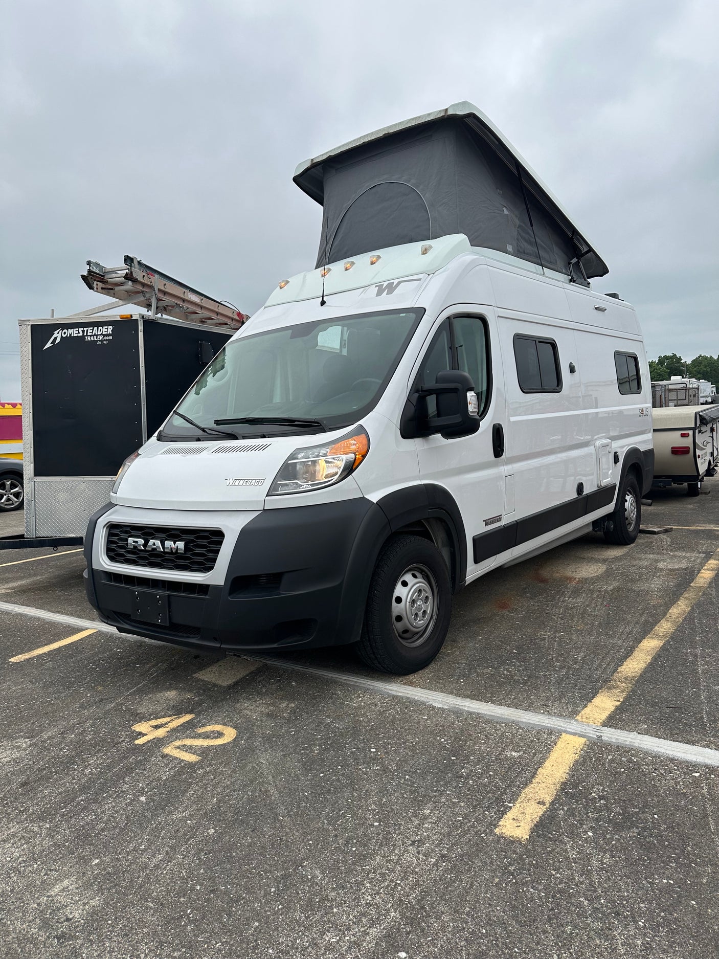 White Winnebago Solis pop-top RV rental (Ram ProMaster camper van) parked in lot 42 on a cloudy day.