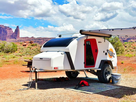 White teardrop RV rental, red door, awning extended, parked on red gravel in a desert with towering rock formations.