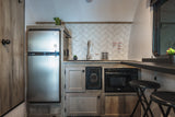 Modern RV rental kitchen showcasing a stainless fridge, sink, microwave, wood cabinets, herringbone backsplash, and a bar.