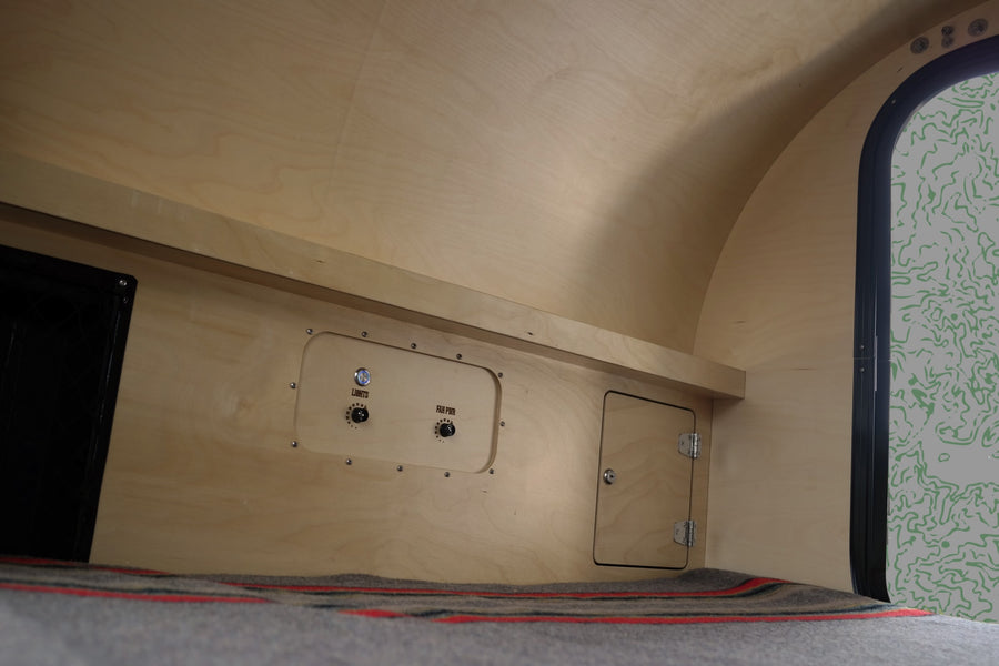 RV rental's plywood interior shows light/fan controls, storage, and a cozy sleeping area with a blanket.