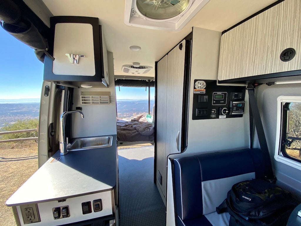 Inside a modern RV rental: kitchen, control panel, and seating with scenic views out the open back doors. Perfect for trips.