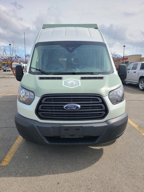 Front of a sage green Ford Transit van, an RV rental, featuring a "VP" logo. Parked outdoors with mountains.