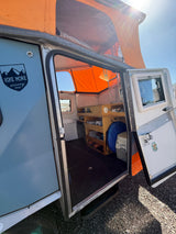 Open door of an RV rental camper reveals a compact interior, kitchen, storage, and pop-top tent ready for adventure.