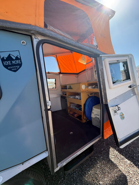 Open door of an RV rental camper reveals a compact interior, kitchen, storage, and pop-top tent ready for adventure.