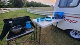 Camping kitchen setup by an RV rental: stove with pot, kettle, table with blue dishes, water jug, and French press.