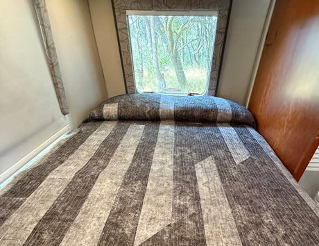 Inside an RV rental: a comfy bed with a patterned quilt and window offering a peaceful forest view, featuring an emergency exit.