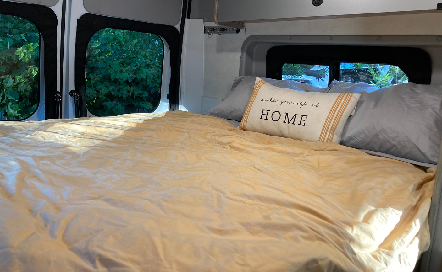 Cozy RV rental bed with "make yourself at HOME" pillow and nature views outside, creating a comfortable travel space.