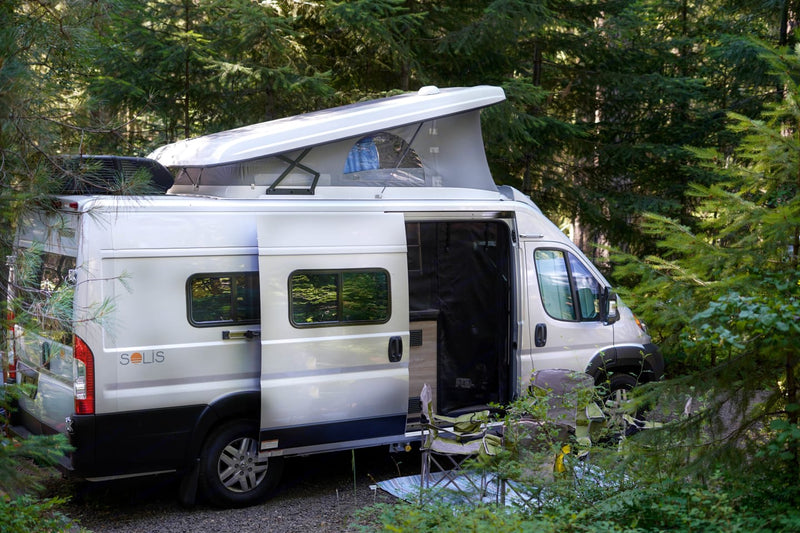  facing a large snow-capped mountain under a clear sky. Explore nature!|A bright Winnebago Solis rv rental van with an open pop-top and side door is parked among green trees — photo 2