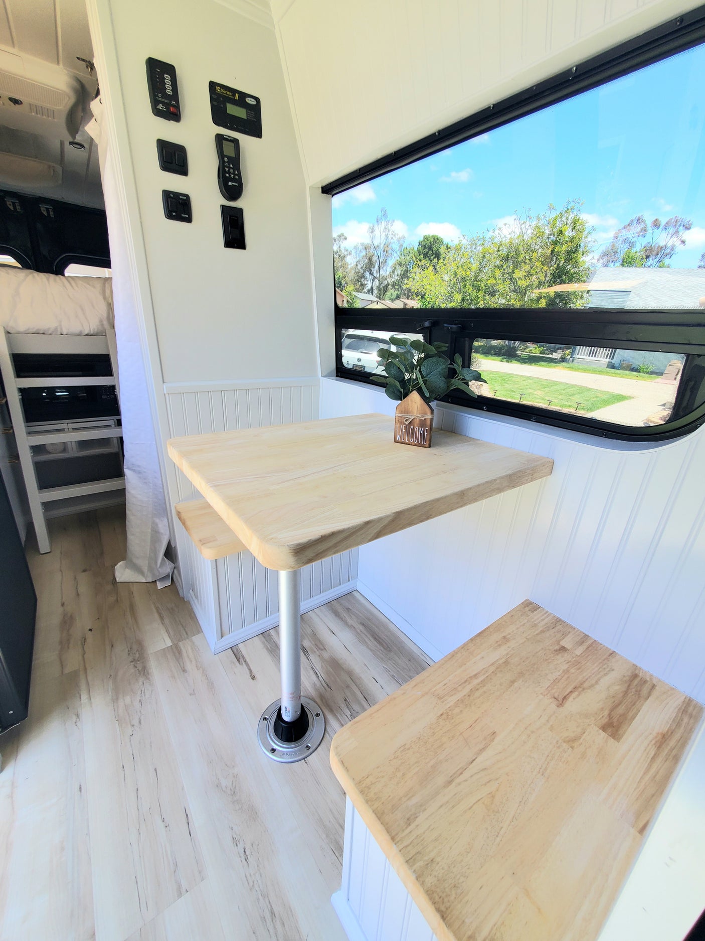 Inside a modern rv rental: bright dinette with wood table and benches, scenic window view of trees, and various control panels.
