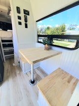 Inside a modern rv rental: bright dinette with wood table and benches, scenic window view of trees, and various control panels.