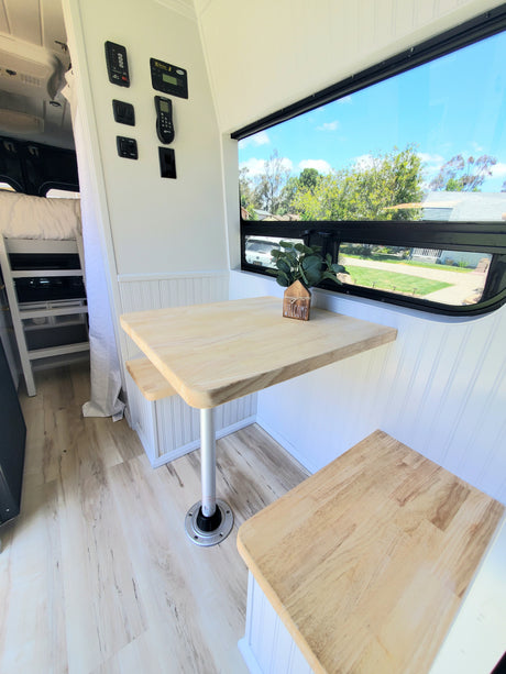 Inside a modern rv rental: bright dinette with wood table and benches, scenic window view of trees, and various control panels.