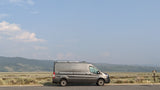 A grey Ford Transit camper van (RV rental) parked at a scenic overlook with distant mountains and a "No Camping" sign.
