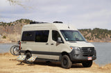 This Geotrek Mercedes-Benz Sprinter camper van, an ideal rv rental, is parked with a bike & chairs by a scenic lake.