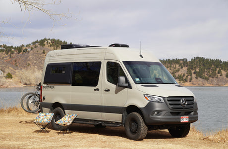 This Geotrek Mercedes-Benz Sprinter camper van, an ideal rv rental, is parked with a bike & chairs by a scenic lake.