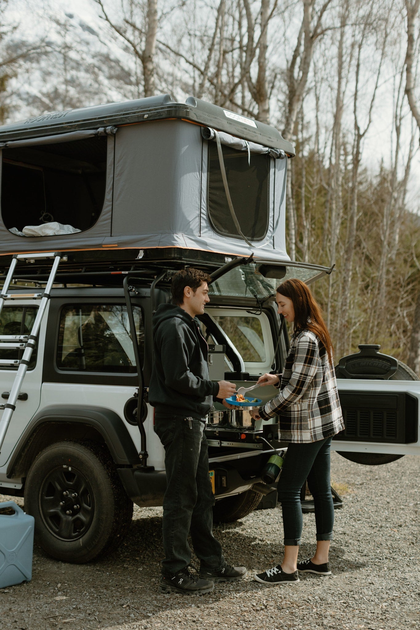 Couple cooks with a Roofnest rooftop tent on their RV rental, a compact camper setup for outdoor adventures.