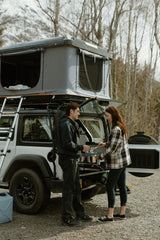 Couple cooks with a Roofnest rooftop tent on their RV rental, a compact camper setup for outdoor adventures.